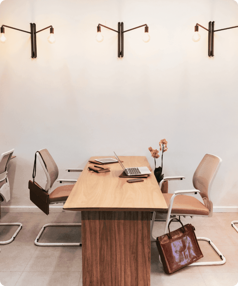 Modern wooden office desk with two chairs, laptops, bags, and orange flowers against white wall with pendant lights.