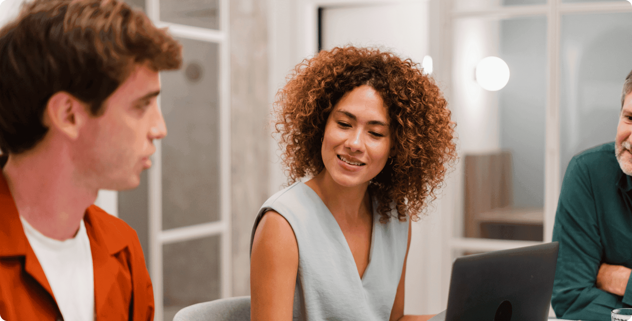 Four people seated at table shaking hands across documents, with large plant in background.