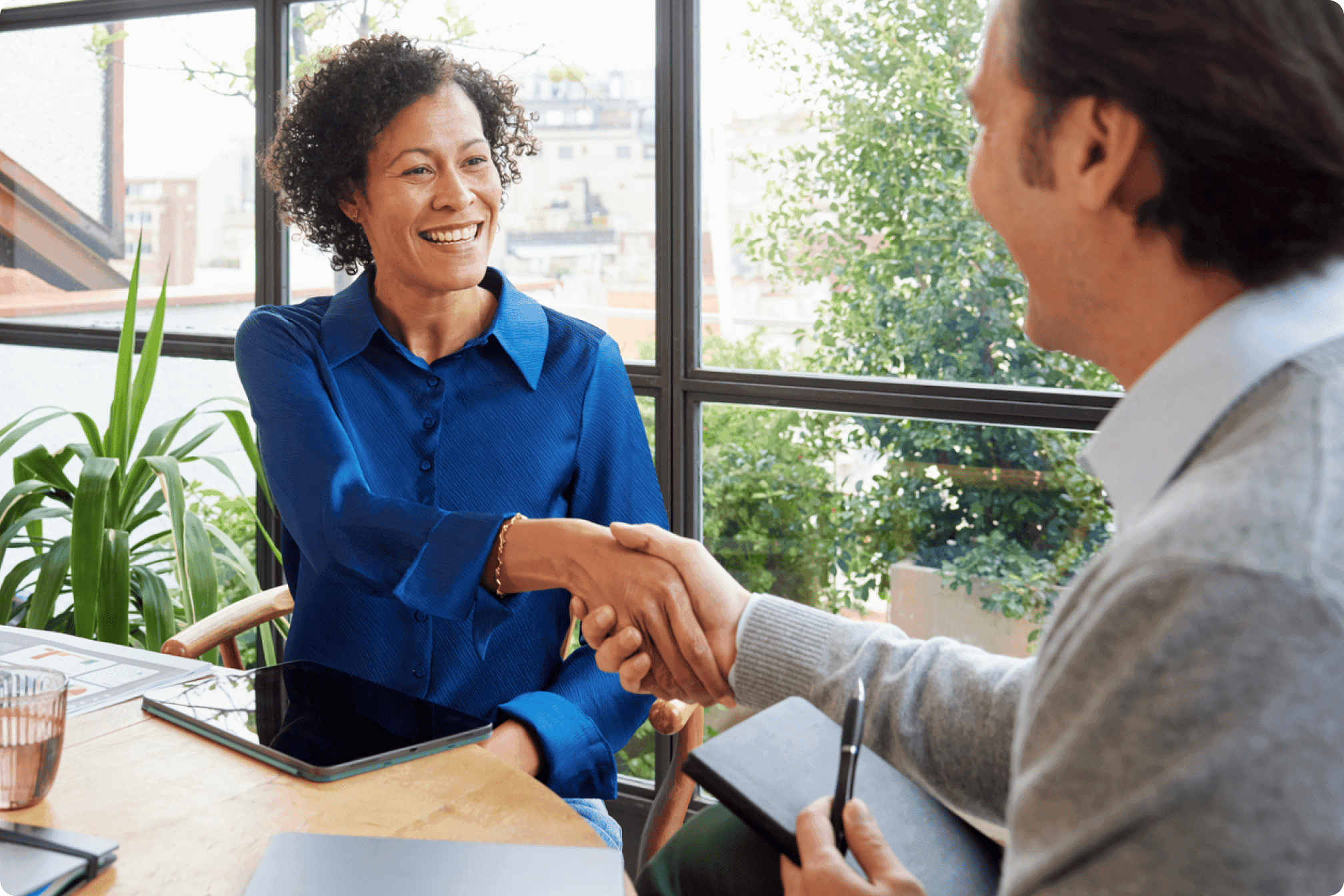 Two professionals sitting by large window shaking hands across a table with tablet and notebook.