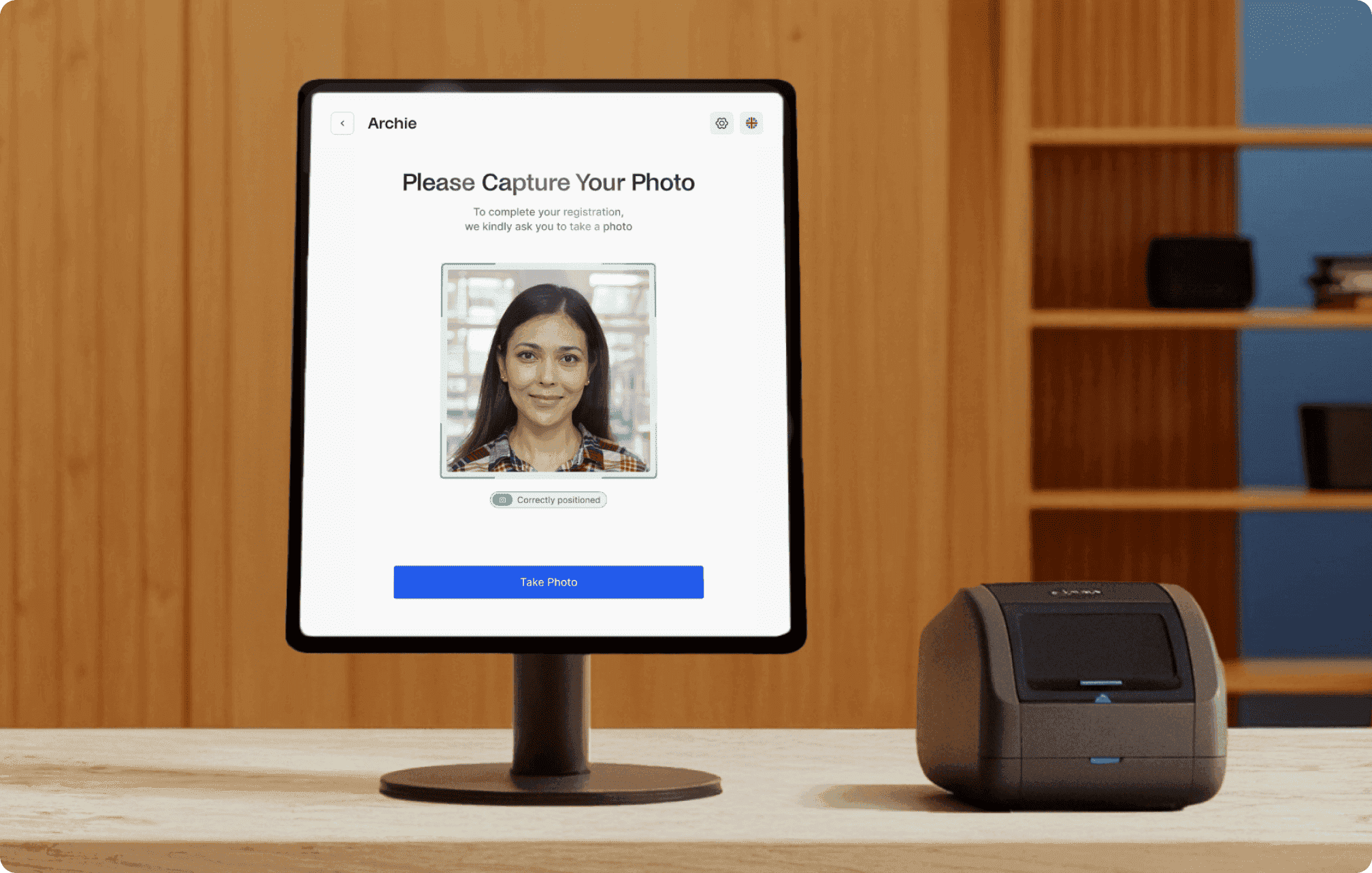 Tablet kiosk on wooden desk displaying “Archie Capture Your Photo” screen with frame for face scan, green checkmark, blue retake button, and ID scanner beside it.