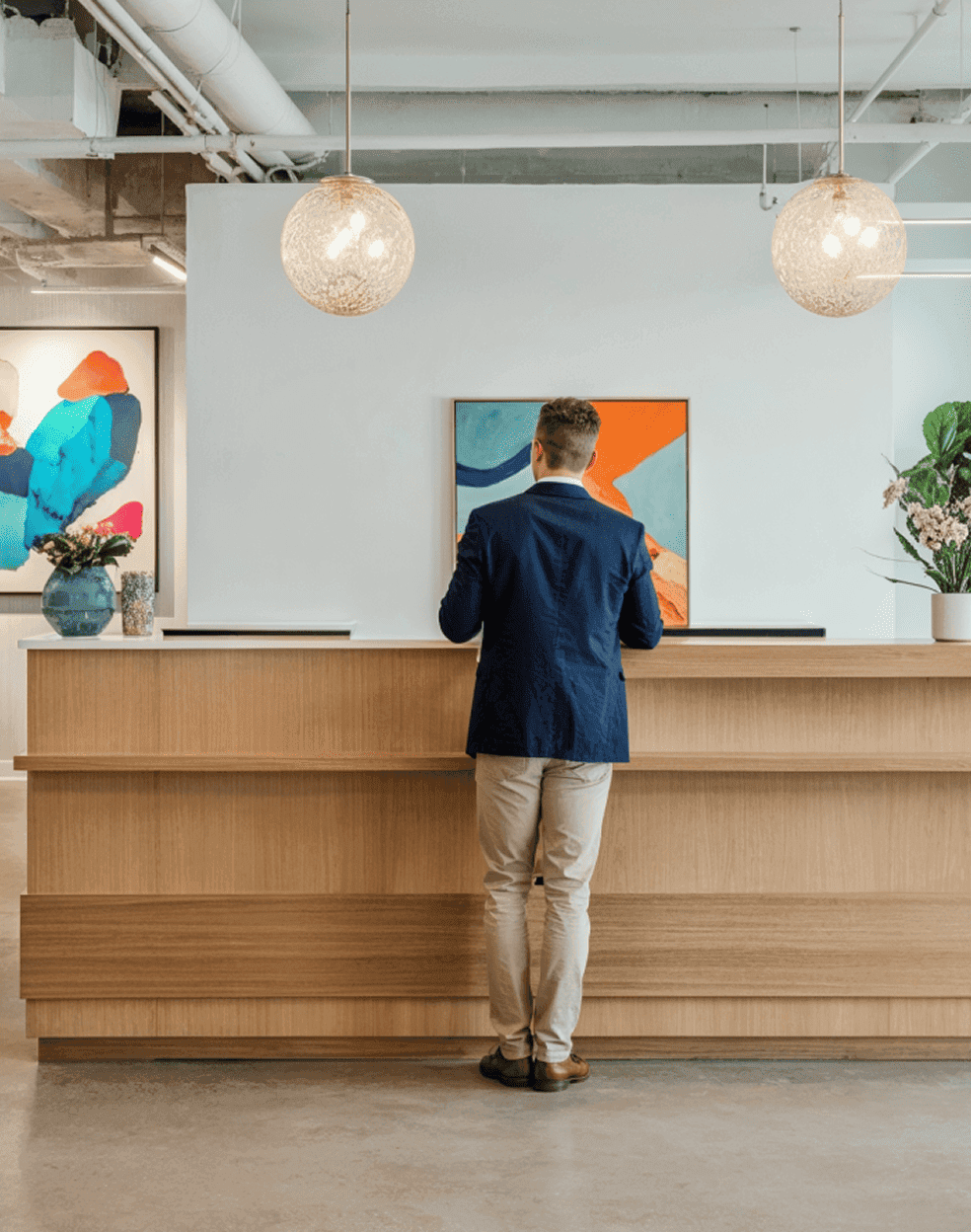Un homme debout à une réception moderne dans un bureau, avec de l'art abstrait et des lampes suspendues en arrière-plan.