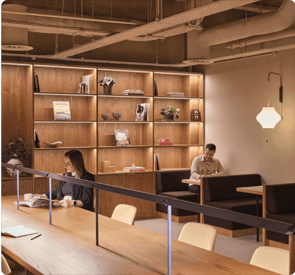 Un espace de co-working moderne avec deux personnes travaillant à une longue table, avec des étagères en bois avec des livres et des objets décoratifs en arrière-plan.
