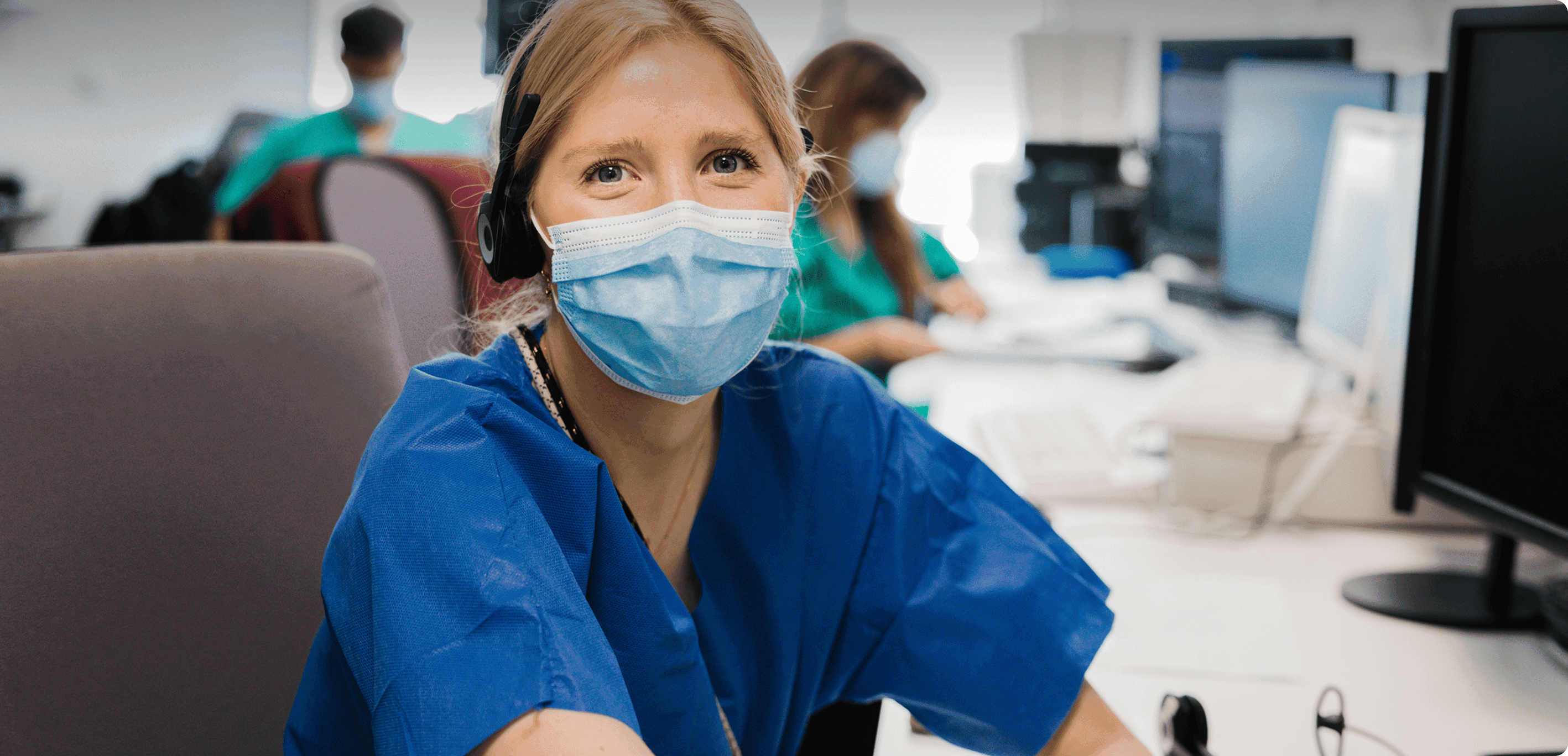 Healthcare worker in blue scrubs at a desk with computer and headset, colleagues working in background
