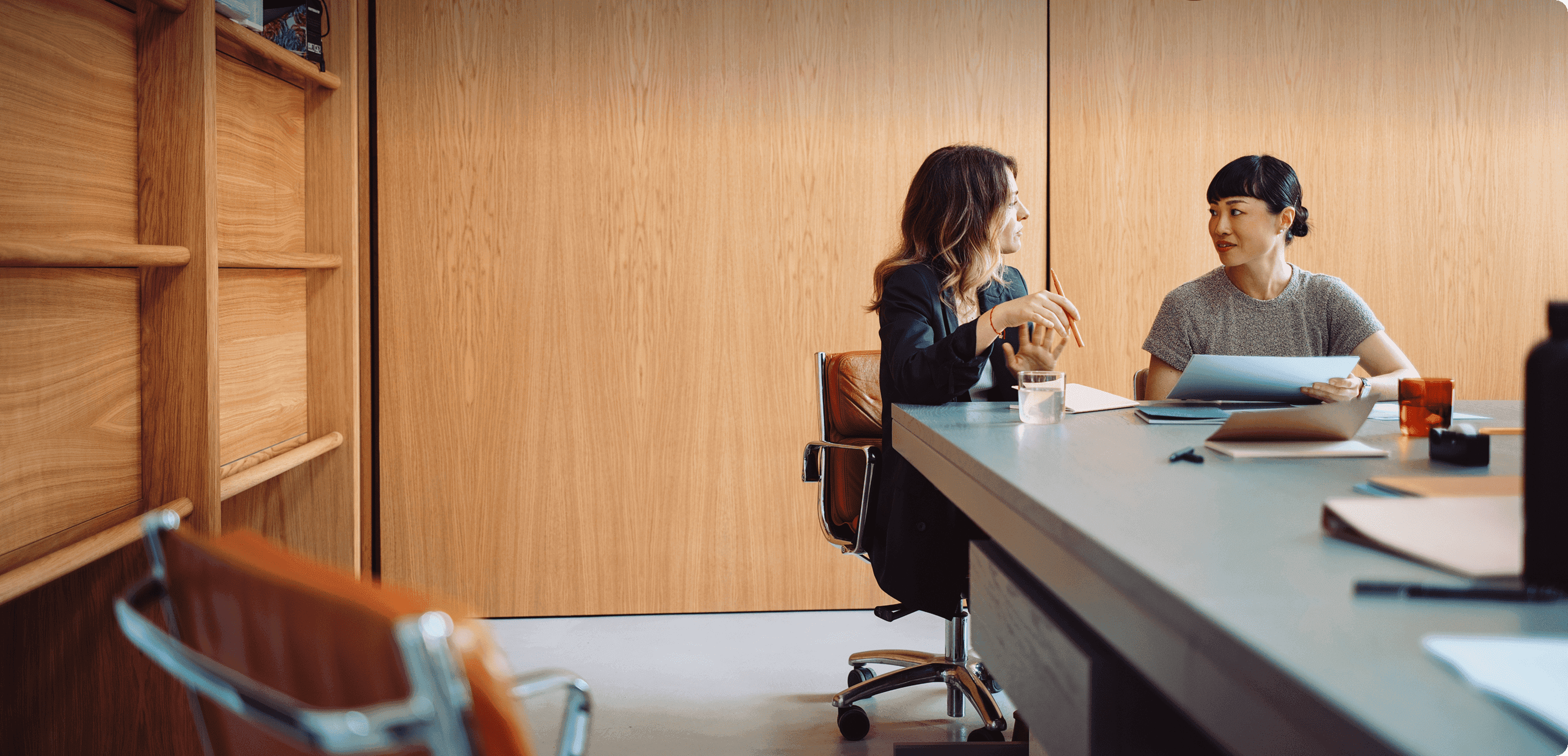 Two women seated at a table in a modern office with wooden paneling, discussing documents