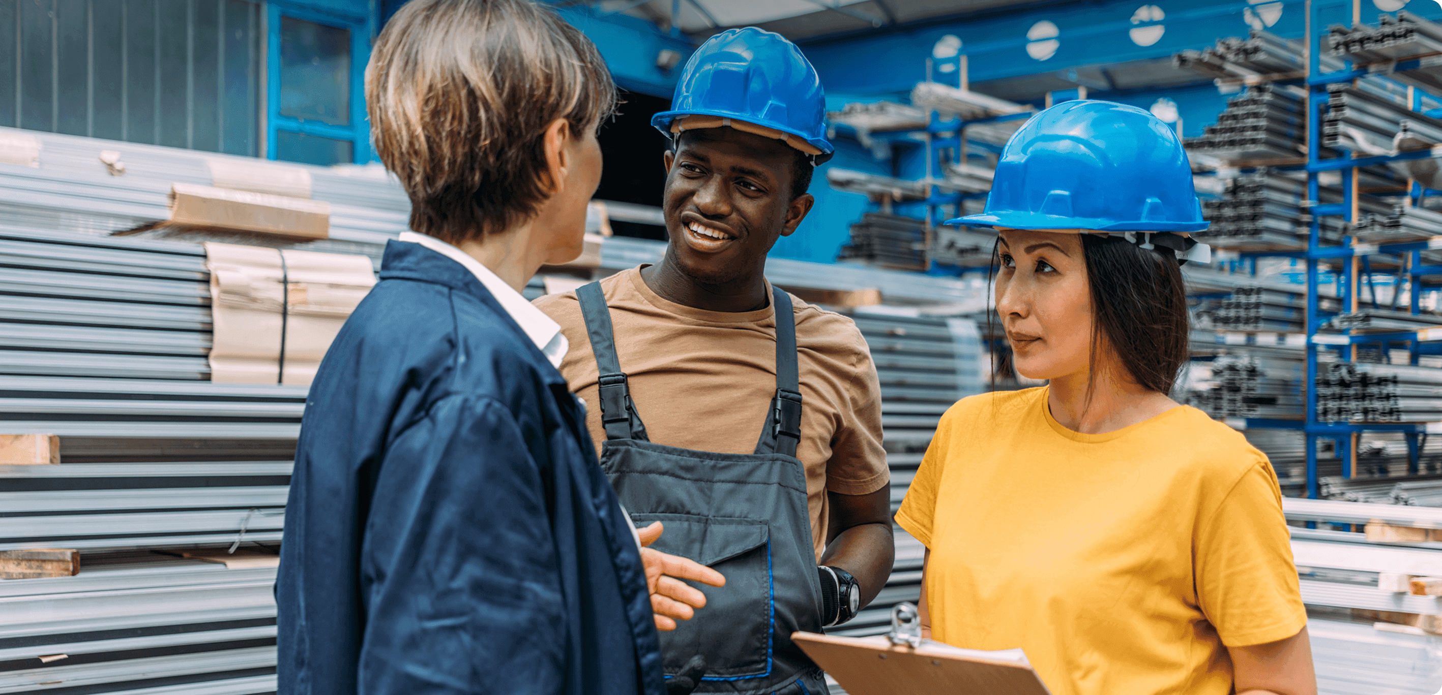 Three people, two in blue hard hats, discuss in front of stacked metal pipes at an industrial site.