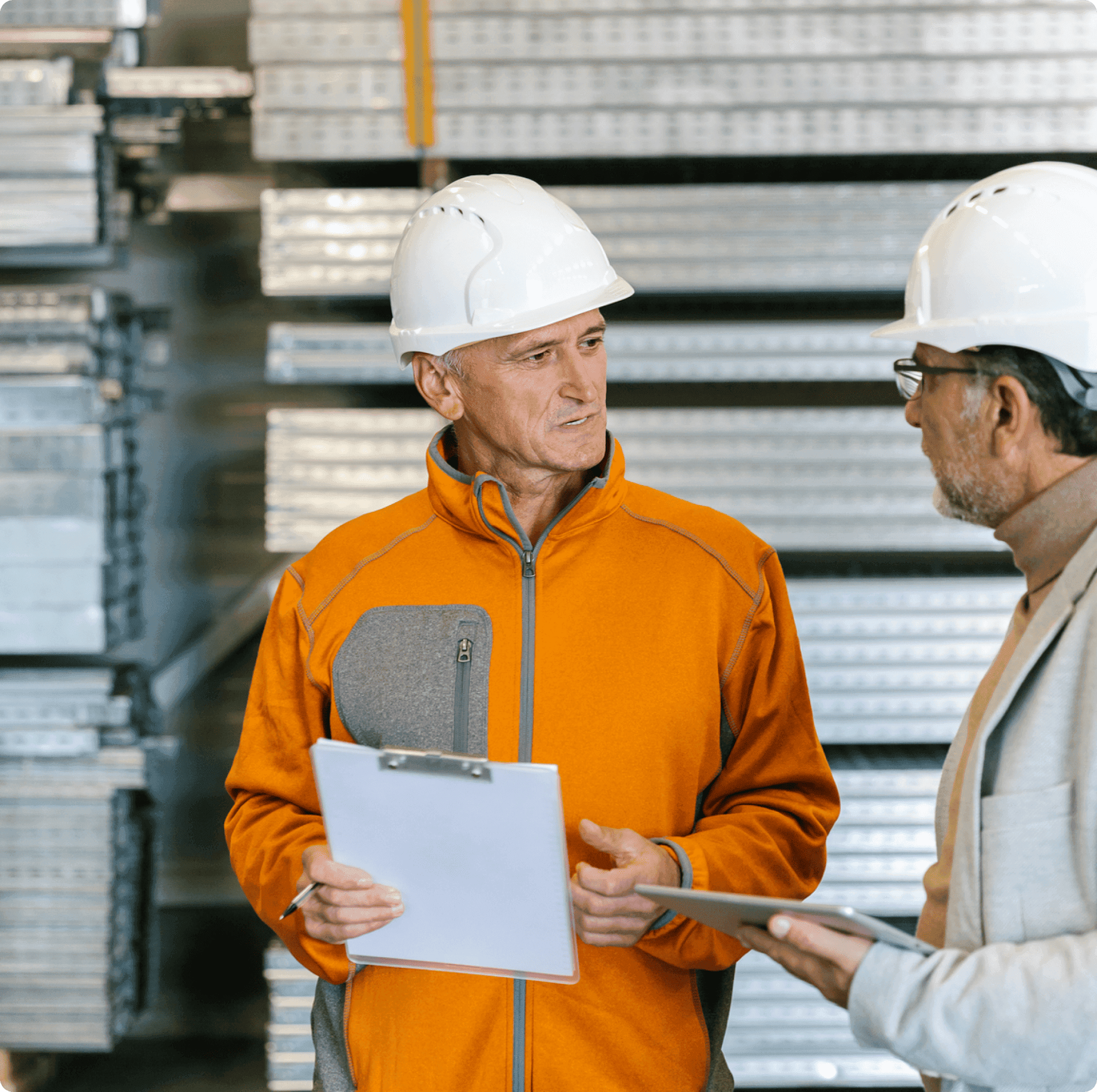 Two people wearing white safety helmets and orange or grey jackets discuss in front of stacked metal parts.
