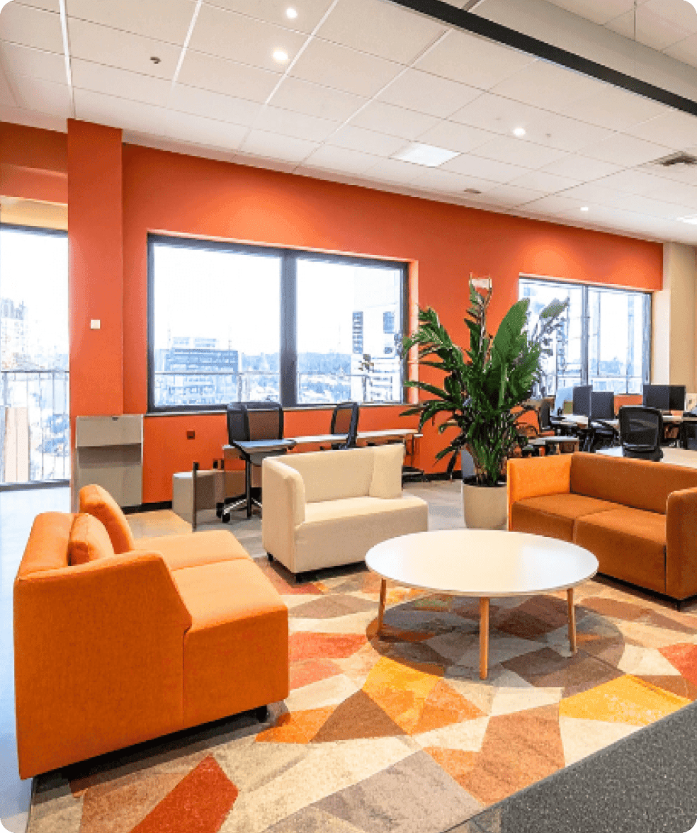Modern office lounge with orange walls, bright windows, orange and beige sofas, and round table.