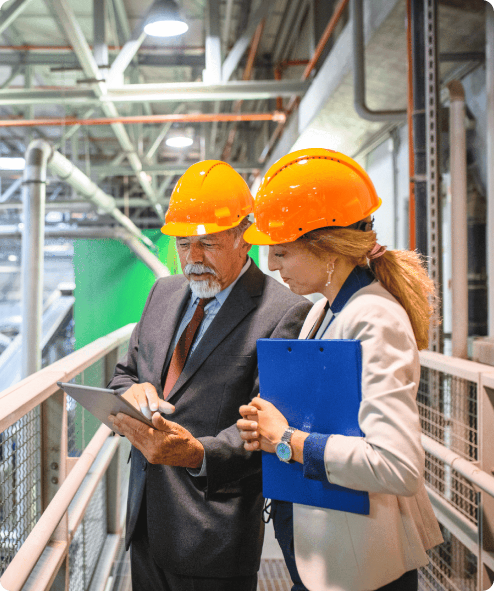 Two people in suits and orange safety helmets consulting over a clipboard on an industrial walkway.
