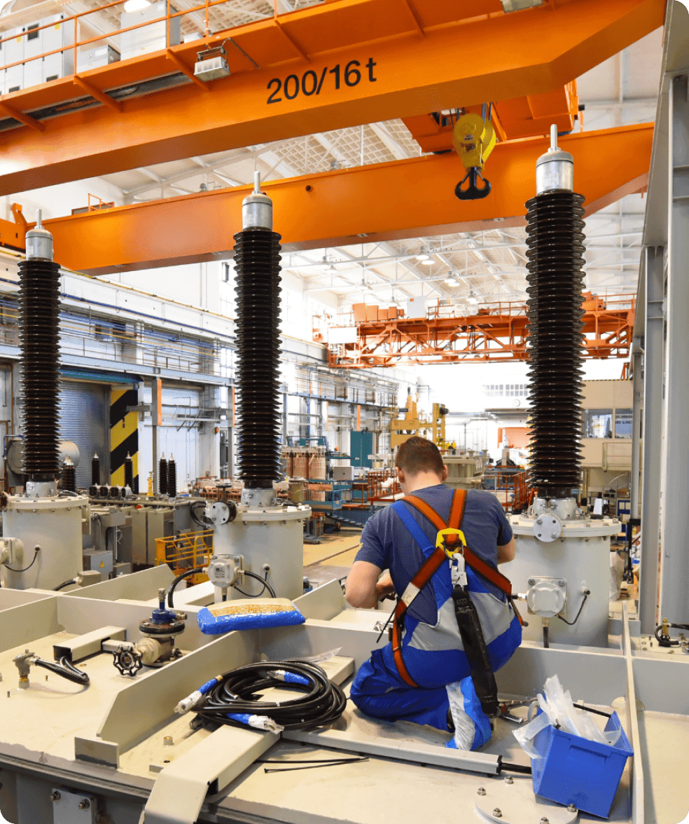 Industrial worker in blue uniform working among large electrical equipment and orange overhead crane.