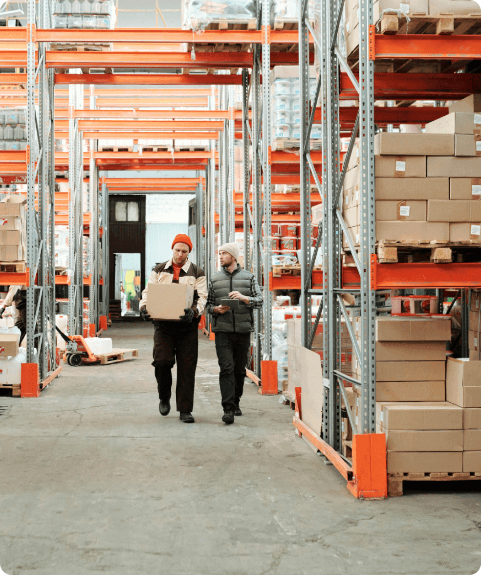 Two people walk through tall warehouse aisles carrying boxes, surrounded by orange shelving.