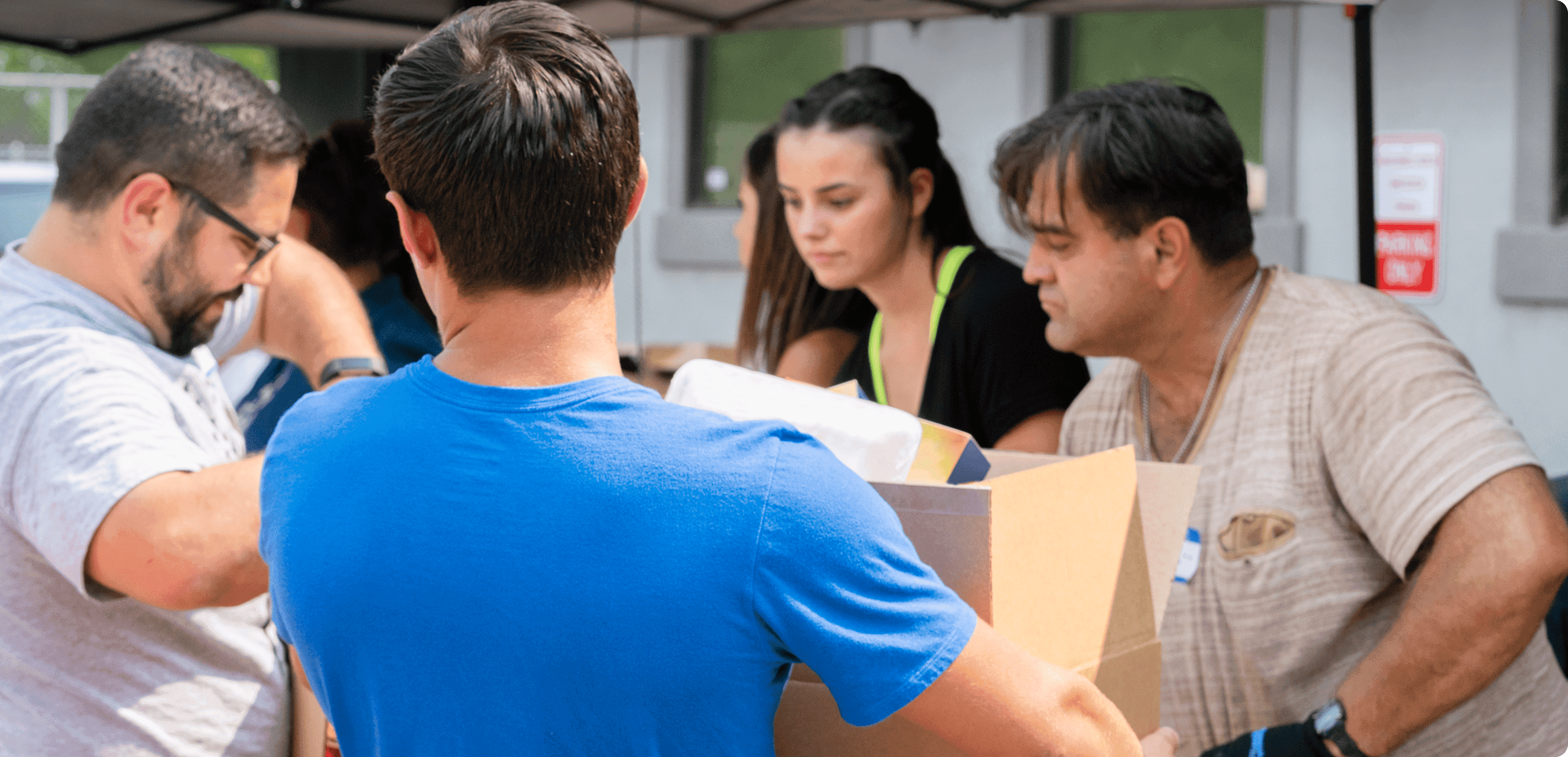 Volunteers interact and sort boxes during an outdoor community event, under tent canopies.