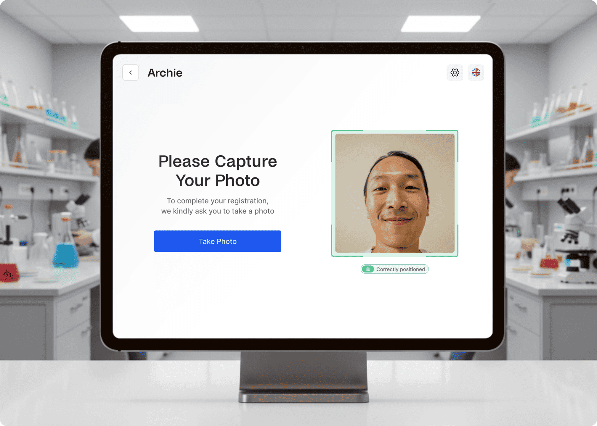 A registration screen prompts a lab visitor to take a photo for entry, with laboratory background visible on the monitor.