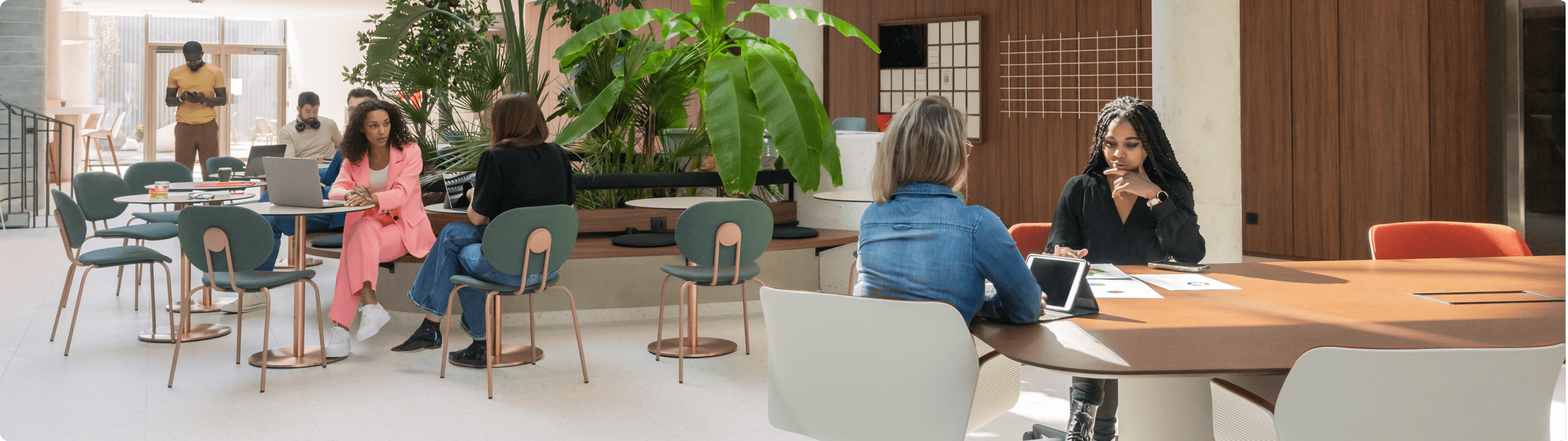 Bright, open plan office lounge area with multiple people seated at tables surrounded by plants