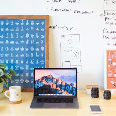 A creative workspace with a laptop on a wooden desk, surrounded by a coffee mug, books, camera lenses, headphones, and framed artwork.