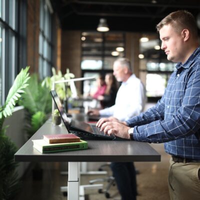 A modern office space with a row of height-adjustable standing desks positioned near large windows that let in natural light.