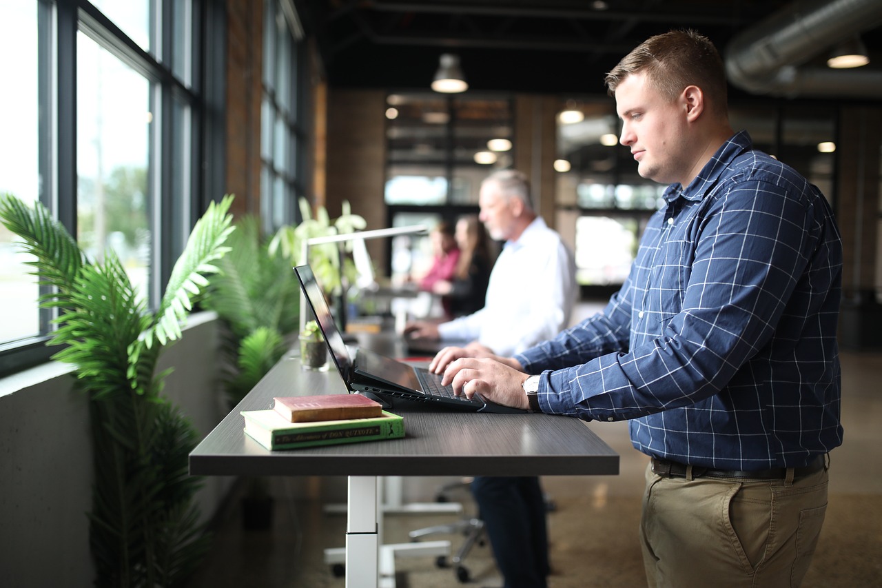 A modern office space with a row of height-adjustable standing desks positioned near large windows that let in natural light.