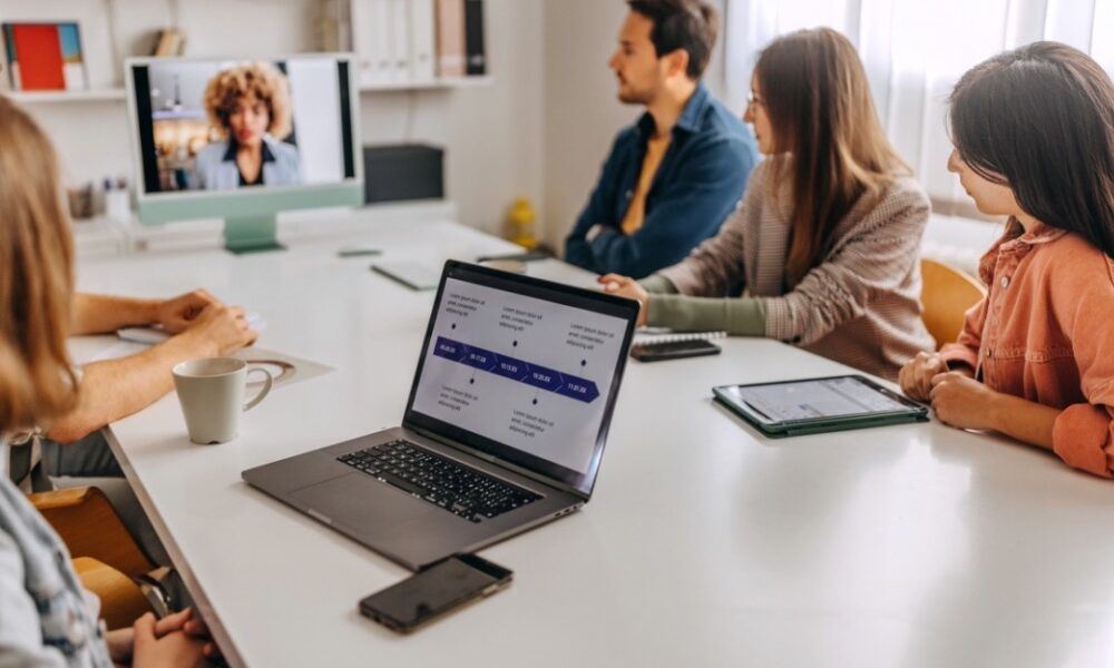 Un grupo de personas en una sala de reuniones sala una videoconferencia con un compañero que aparece en una pantalla.