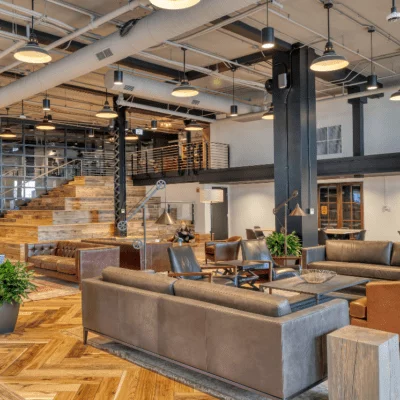 Modern coworking space with leather sofas, wooden floors, open seating areas, and a large staircase leading to an upper level.