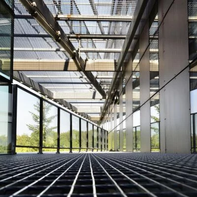 A modern glass walkway with a metal grid floor and overhead metal beams, surrounded by large windows and greenery outside.