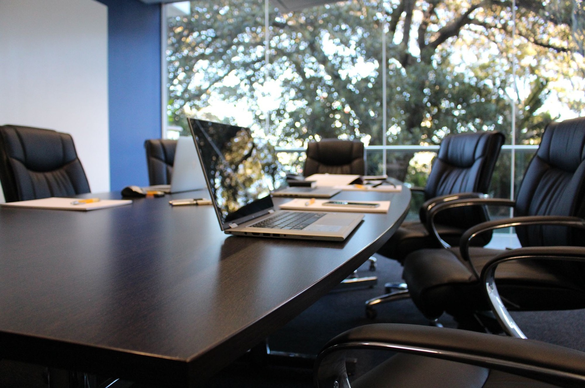 A modern conference room with a large, dark wooden oval table surrounded by black leather chairs with armrests.