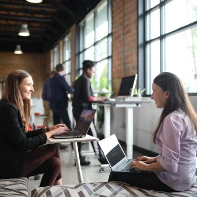 Two women working on laptops and smiling in a modern office space with large windows and a relaxed atmosphere.