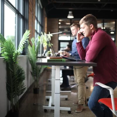 Two men working at adjustable standing desks positioned near large windows that let in ample natural light.