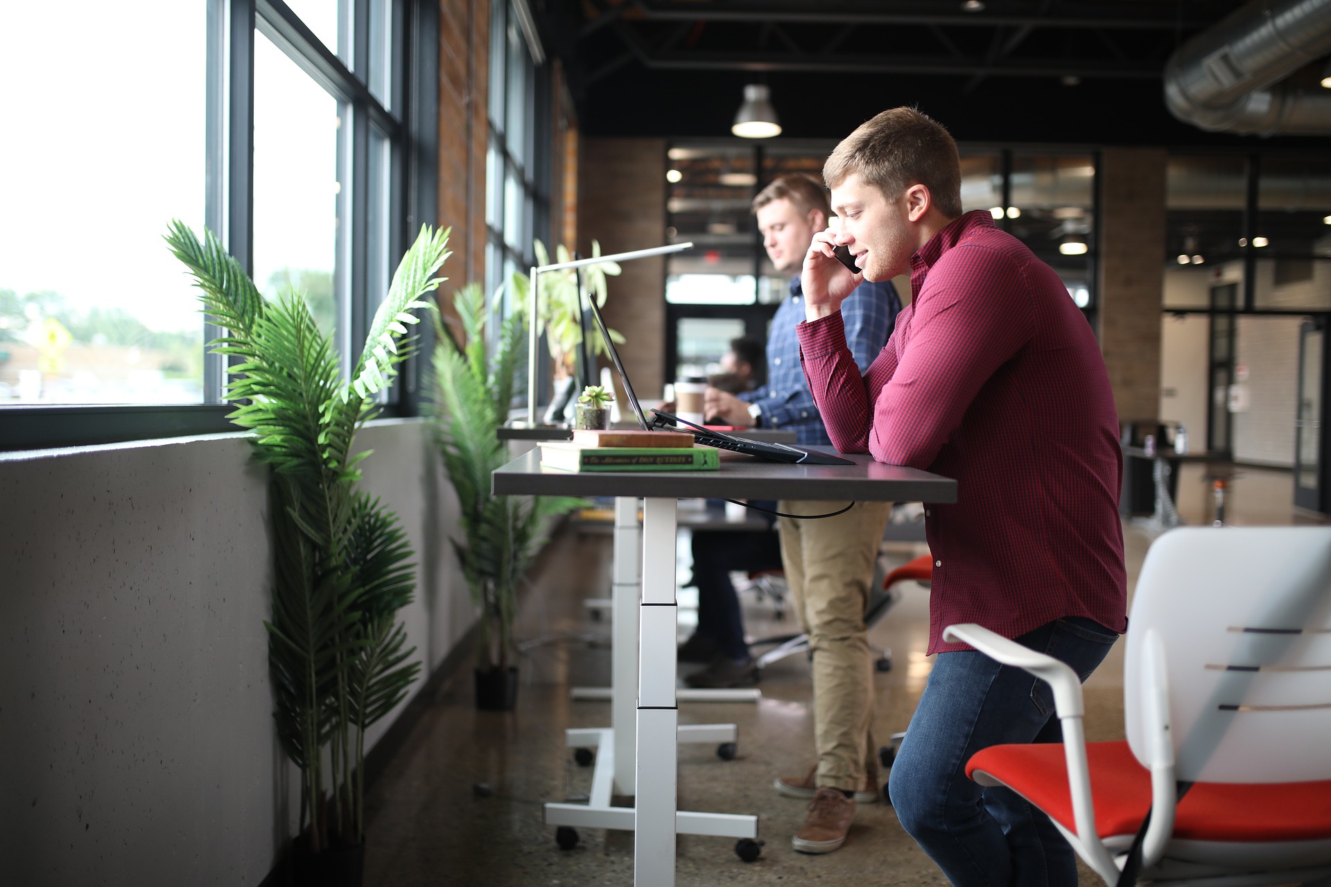 Two men working at adjustable standing desks positioned near large windows that let in ample natural light.