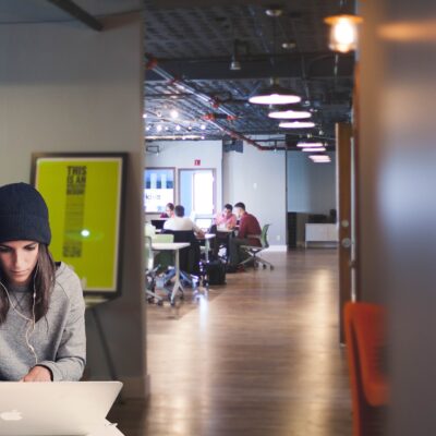 A modern coworking space with a young woman sitting in the foreground, focused on her laptop.