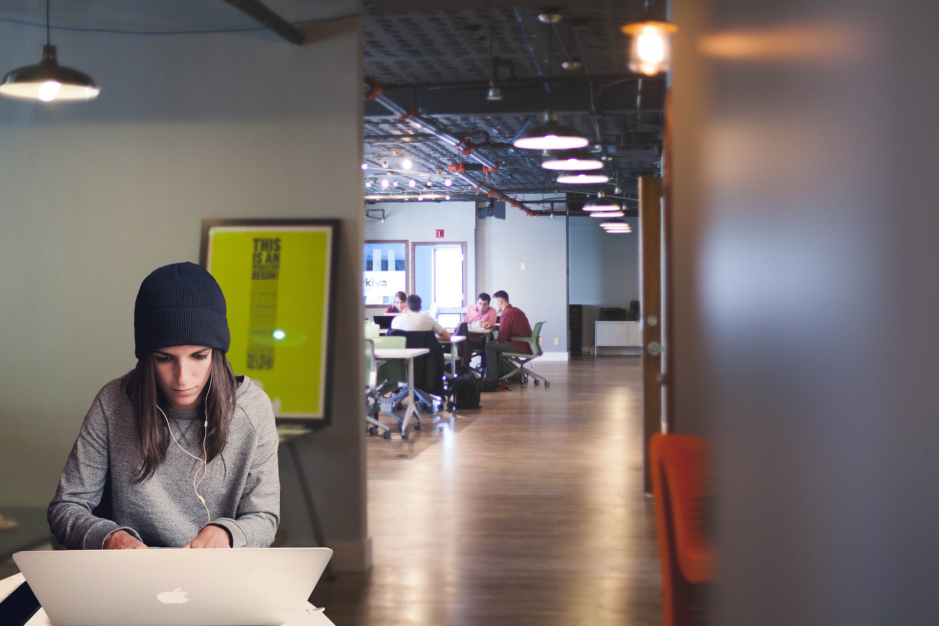 A modern coworking space with a young woman sitting in the foreground, focused on her laptop.