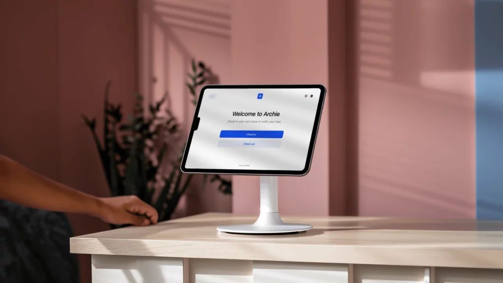A sleek tablet with Archie Visitors app, mounted on a modern white stand, positioned on a light wooden counter.
