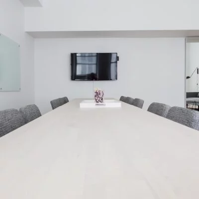 A modern conference room with a long wooden table, gray upholstered chairs, a wall-mounted TV, and glass whiteboards.