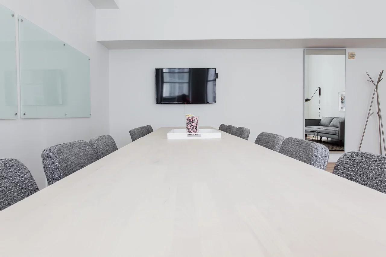 A modern conference room with a long wooden table, gray upholstered chairs, a wall-mounted TV, and glass whiteboards.