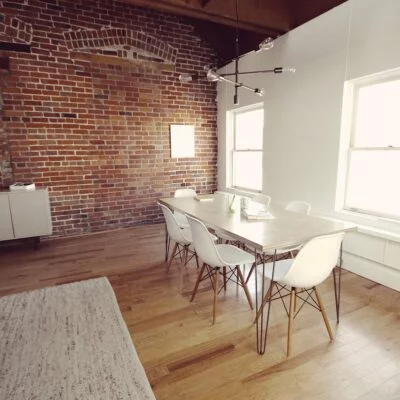 A modern meeting room with a wooden table, white chairs, exposed brick wall, and large windows letting in natural light.