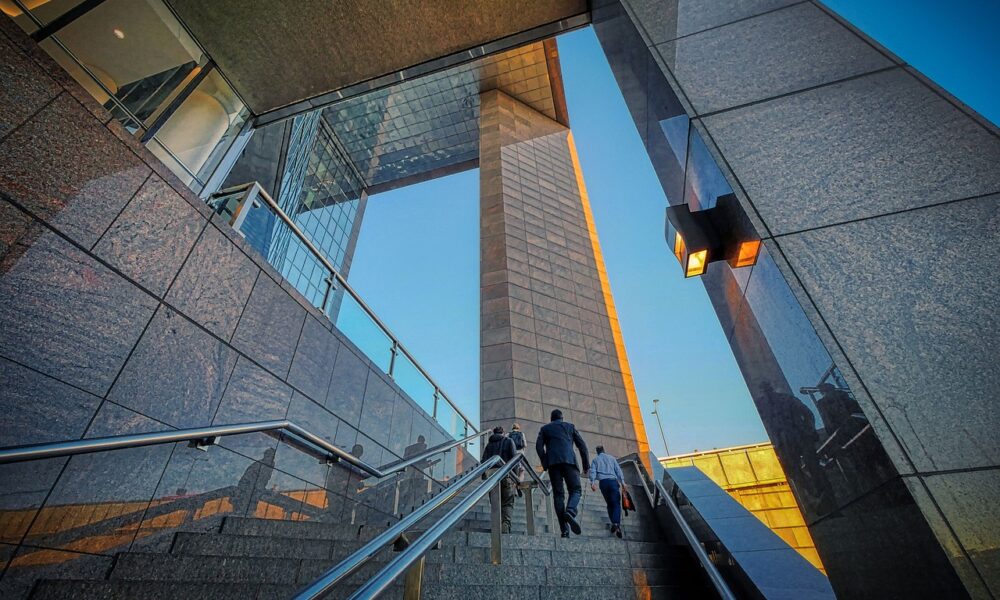 Personas subiendo una escalera rodeada de una arquitectura moderna de cristal y piedra, que refleja la luz del sol del atardecer.