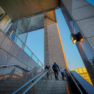 People walking up a staircase surrounded by modern glass and stone architecture, reflecting the evening sunlight.