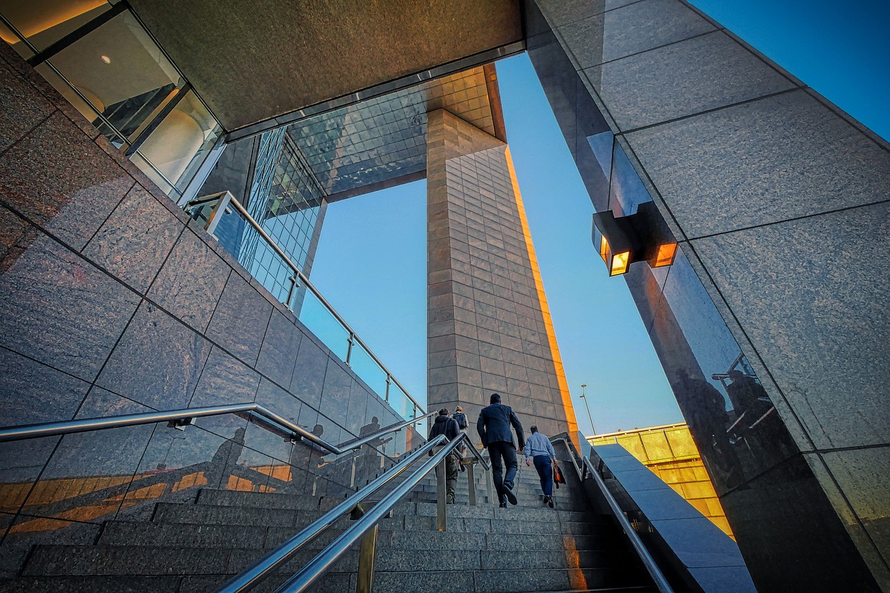 People walking up a staircase surrounded by modern glass and stone architecture, reflecting the evening sunlight.