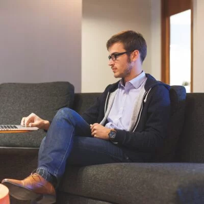 A young man in glasses and business casual attire sits on a dark couch, working on his laptop placed on a small orange side table.