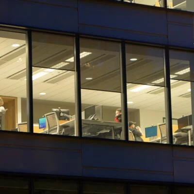 An office building at night with large windows showing people working at desks with computers.