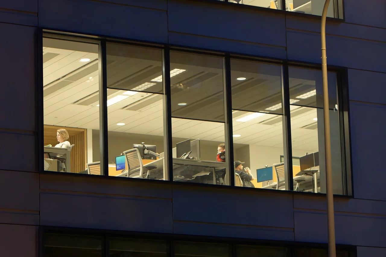 An office building at night with large windows showing people working at desks with computers.