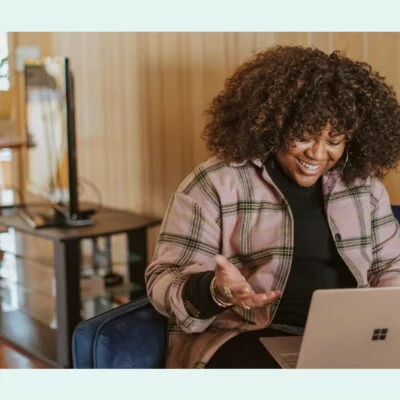 Smiling woman sitting on a couch, video chatting on her laptop in a cozy, wood-paneled room.