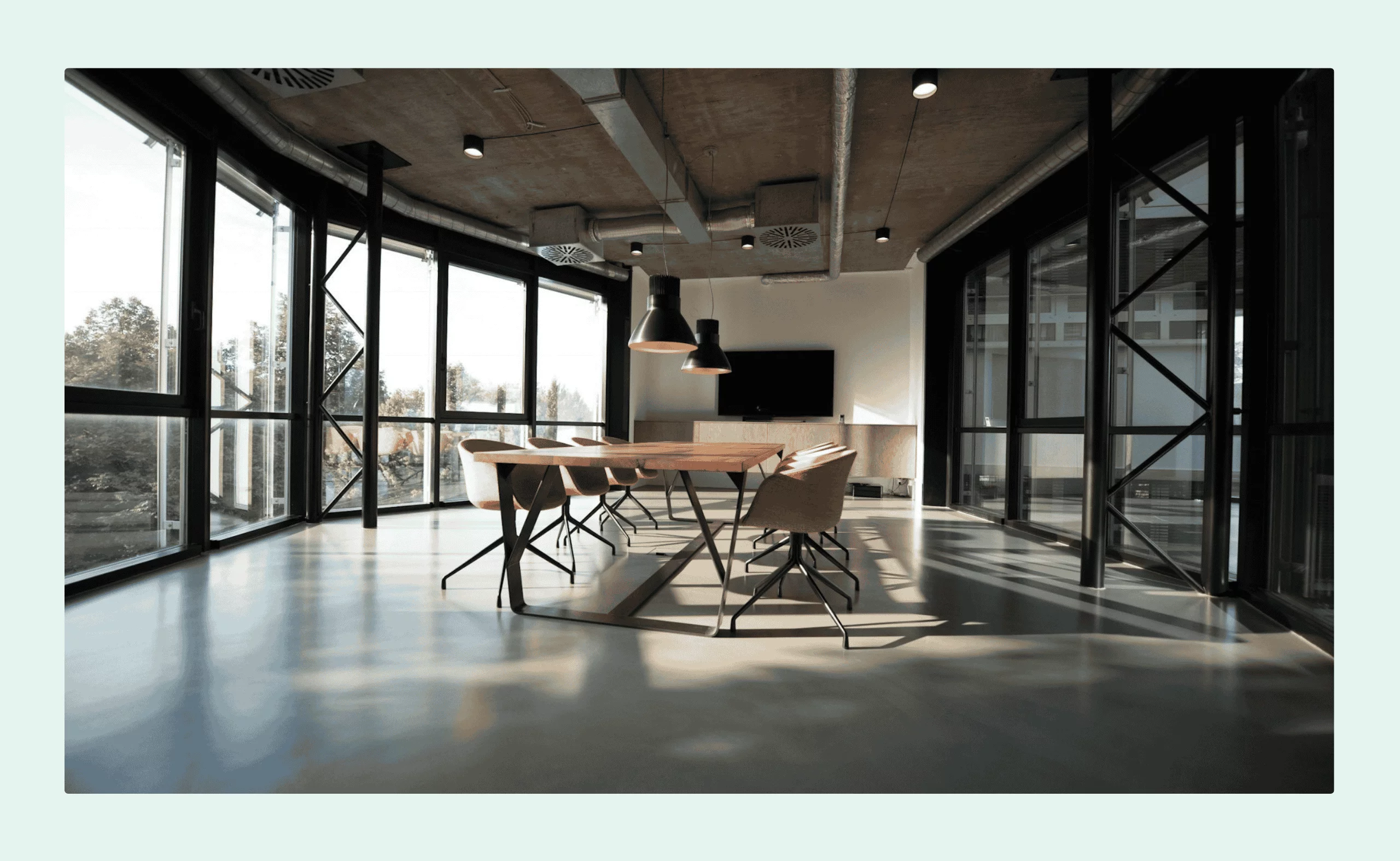 Modern meeting room with large windows, a wooden conference table, pink chairs, and industrial-style ceiling lighting.