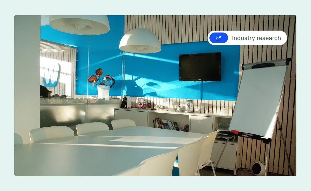 A bright meeting room with a large table, white chairs, a TV on the wall, and a flip chart stand.