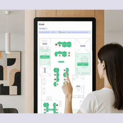 50+ Best (Hot) Desk Booking Software Compared: 2025 Guide A woman using a touchscreen kiosk to view a digital office floor plan and desk availability.