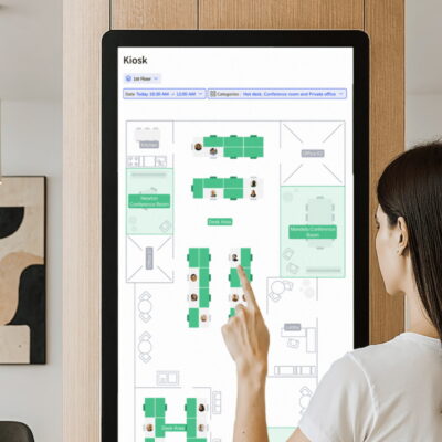 A woman using a touchscreen kiosk to view a digital office floor plan and desk availability.