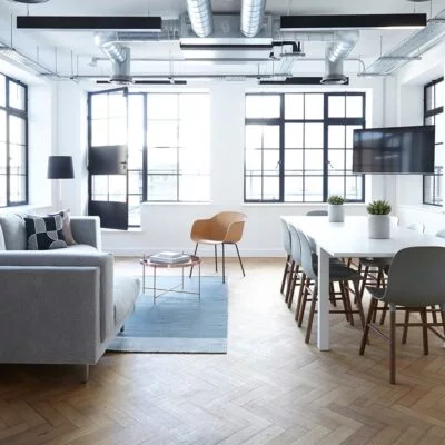 Modern office lounge with a grey sofa, white meeting table, and large windows letting in natural light.
