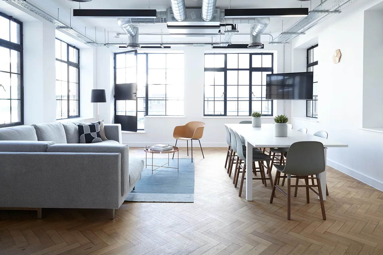 Modern office lounge with a grey sofa, white meeting table, and large windows letting in natural light.