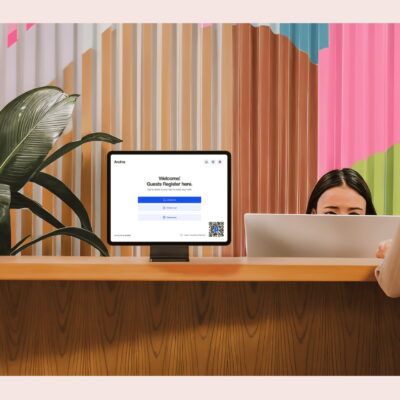 A woman checks in at a modern office reception desk with a visitor registration screen, while a receptionist sits behind a computer.