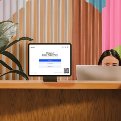 A woman checks in at a modern office reception desk with a visitor registration screen, while a receptionist sits behind a computer.