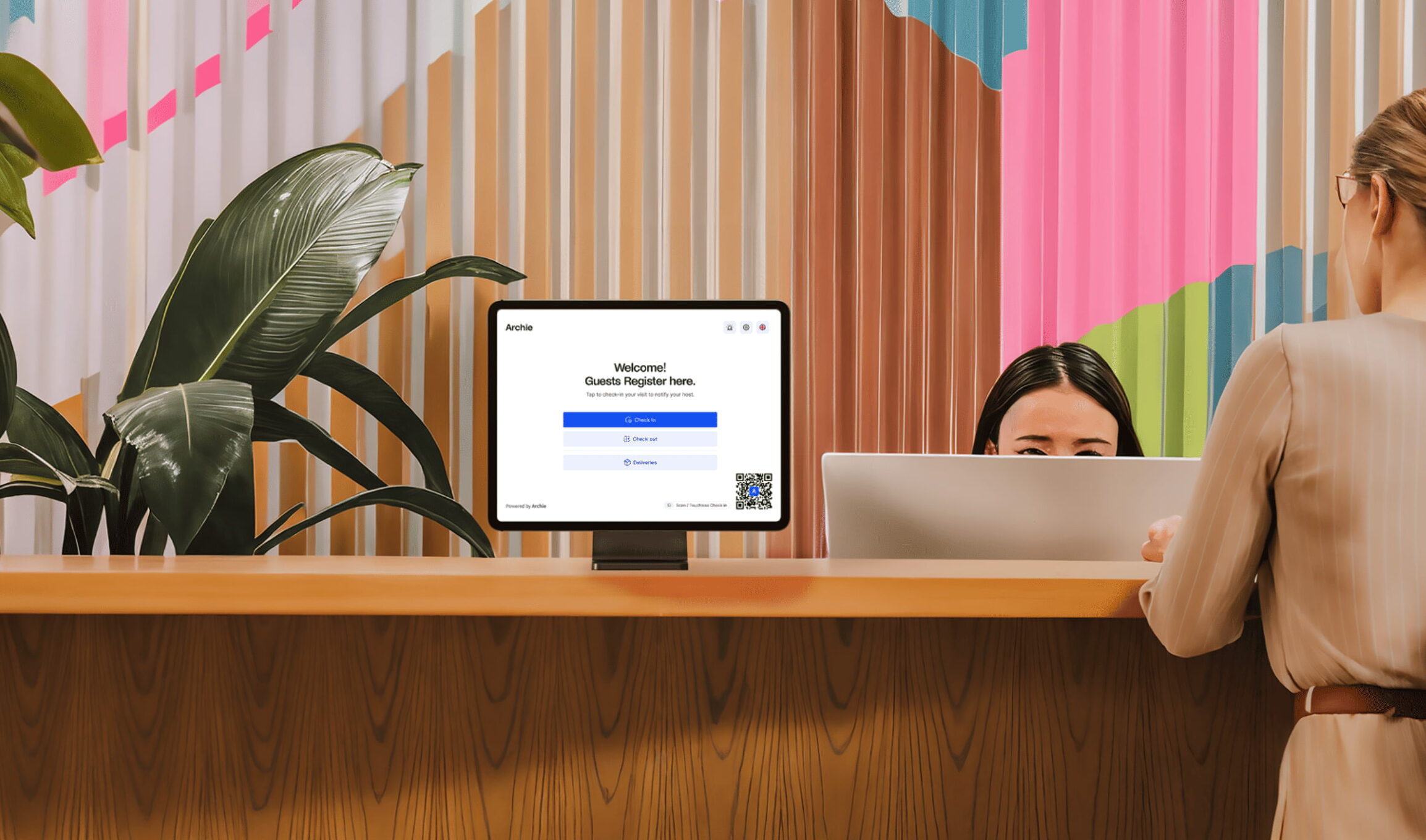 A woman checks in at a modern office reception desk with a visitor registration screen, while a receptionist sits behind a computer.