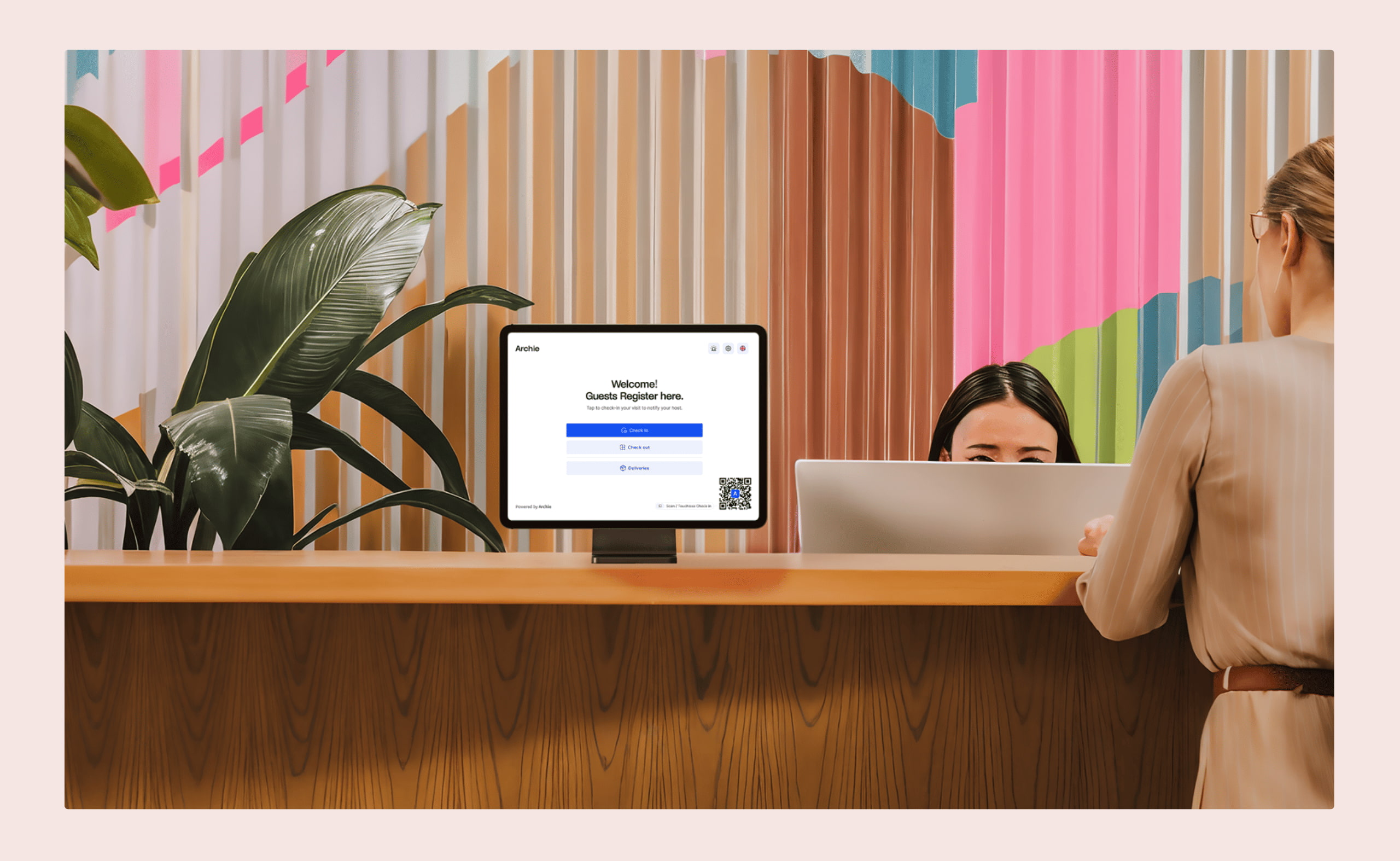A woman checks in at a modern office reception desk with a visitor registration screen, while a receptionist sits behind a computer.